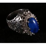 White metal openwork ring set with central blue cabochon encircled by a band of diamonds,