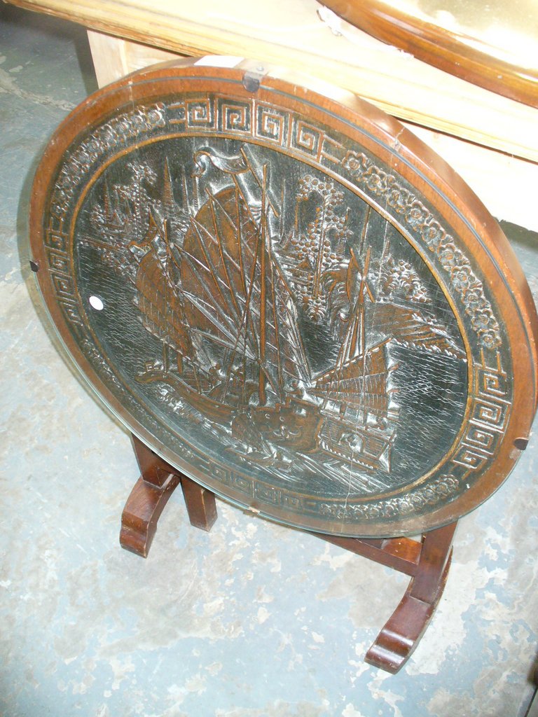 A Japanese folding hardwood circular table carved in deep relief with a masted vessel.