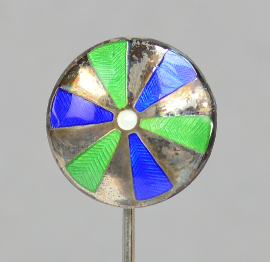 A sterling silver hat pin of circular form with spokes of green and blue enamel radiating from the