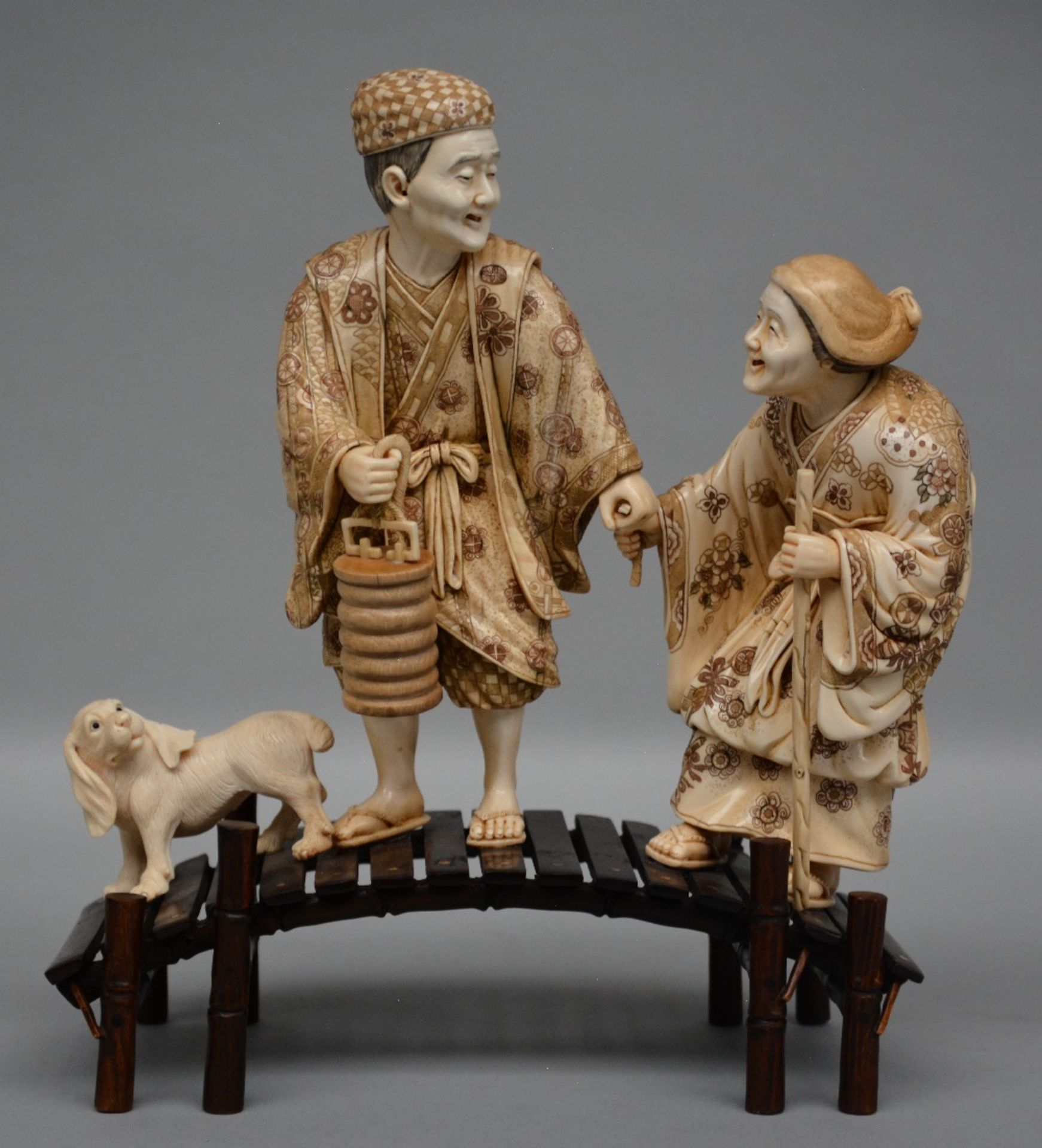 A charming Japanese ivory okimono depicting two figures and a dog on a bridge, red and black