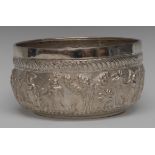 A large Burmese silver coloured metal circular bowl, chased with figures in a village,