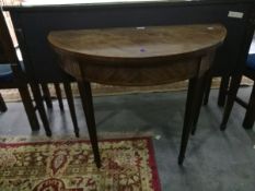 A figured mahogany demi-lune card table on reeded tapering supports (the back leg is coming away,