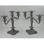 A pair of silver plated three light candelabras, having scrolling end arms and foliate square bases,