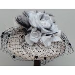 A 'Royal Ascot' labelled cream hat with white flowers and trimmed black feathers arranged over a