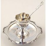 An Edwardian silver egg cup on stand, 15cm wide, with a shaped line border broken by scrolls, with