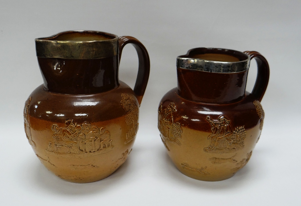TWO DOULTON LAMBETH STONEWARE TAVERN JUGS decorated in relief and with silver collars to both,