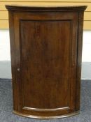 A BOW FRONT OAK HANGING CORNER CUPBOARD having a single inverted panelled door, hinging to reveal