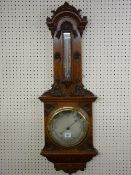 A CARVED OAK ANEROID WALL BAROMETER, elaborately framed with silvered dial having a brass bezel, a