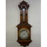 A CARVED OAK ANEROID WALL BAROMETER, elaborately framed with silvered dial having a brass bezel, a