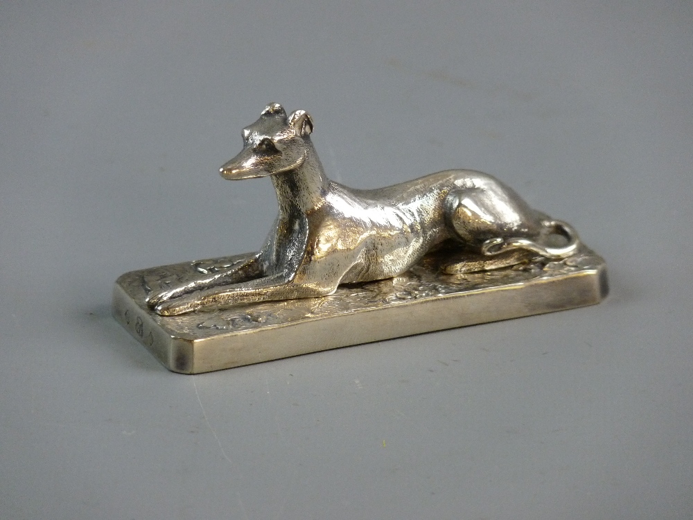 A WHITE METAL FIGURE OF A RECUMBENT GREYHOUND set upon a rectangular plinth with surrounding