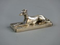 A WHITE METAL FIGURE OF A RECUMBENT GREYHOUND set upon a rectangular plinth with surrounding