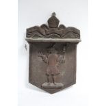 A carved wooden shield-shaped wall plaque depicting a mythical pig character figure & inscribed “