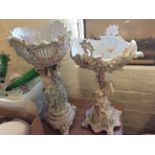 Two ceramic display vases on stands with decorative flowers and figurines.