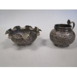 An Indian white metal cream jug and sugar bowl; the jug with a Cobra handle, profusely decorated