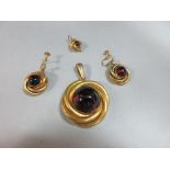 A garnet pendant and earring suite, each with a round cabochon garnet in a closed back collet within