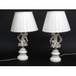 A pair of handed white ceramic table lamps with silk shades, in the form of cherubs with scarves.