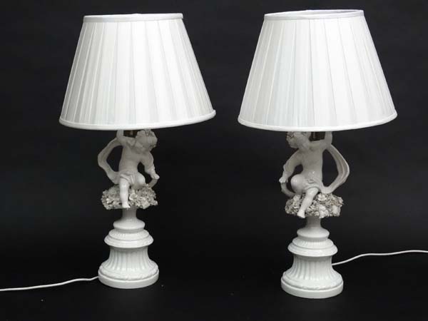 A pair of handed white ceramic table lamps with silk shades, in the form of cherubs with scarves.