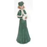 A figure of a lady in green winter attire with white fur trim and applied holly decoration,