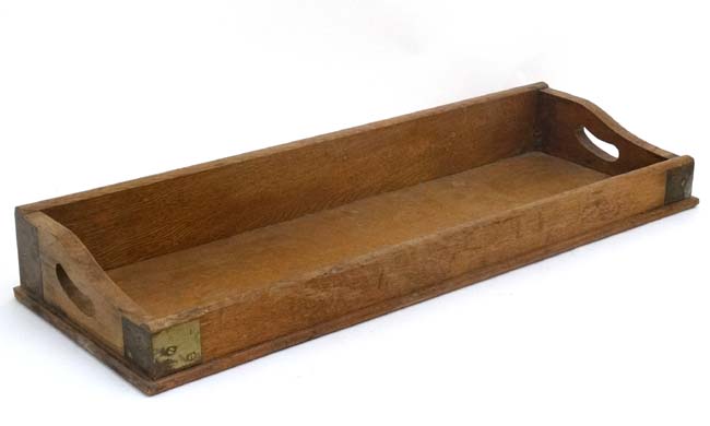 An early 20thC blonde oak desk top book trough / tray with two pierced carry handles and brass