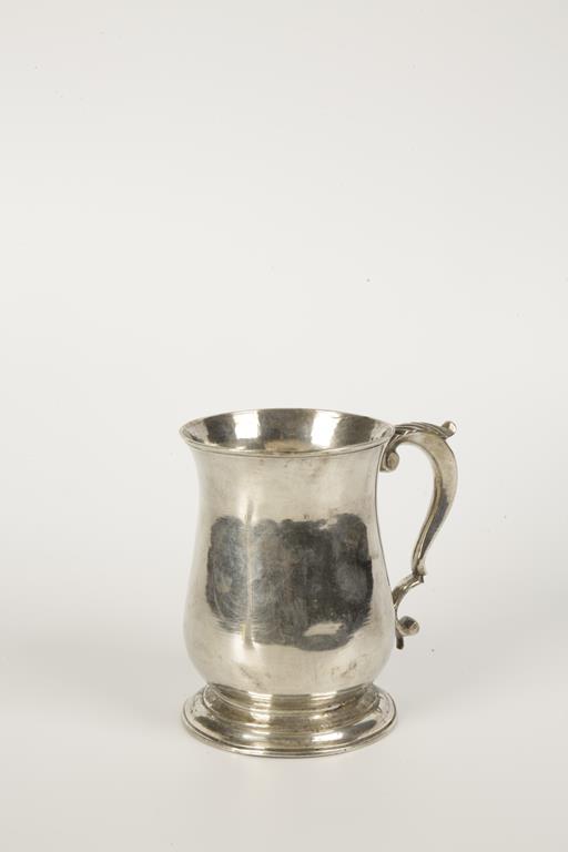 A GEORGE II MUG of baluster form with a leaf capped scroll handle and spreading circular foot,