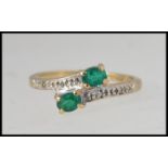 A 9ct gold cross over ring set with two oval cut emeralds and illusion set diamonds.