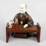 A Japanese Meiji period ivory and lacquer figure of a scholar knelt at a table, with right arm