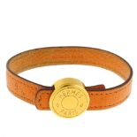 HERMÈS - a bracelet. Designed as an orange leather band to the circular gold coloured clasp which is