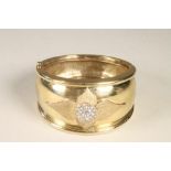 A 9CT GOLD STIFF HINGED BANGLE, the wide convex upper section gypsy set with a roundel of nineteen