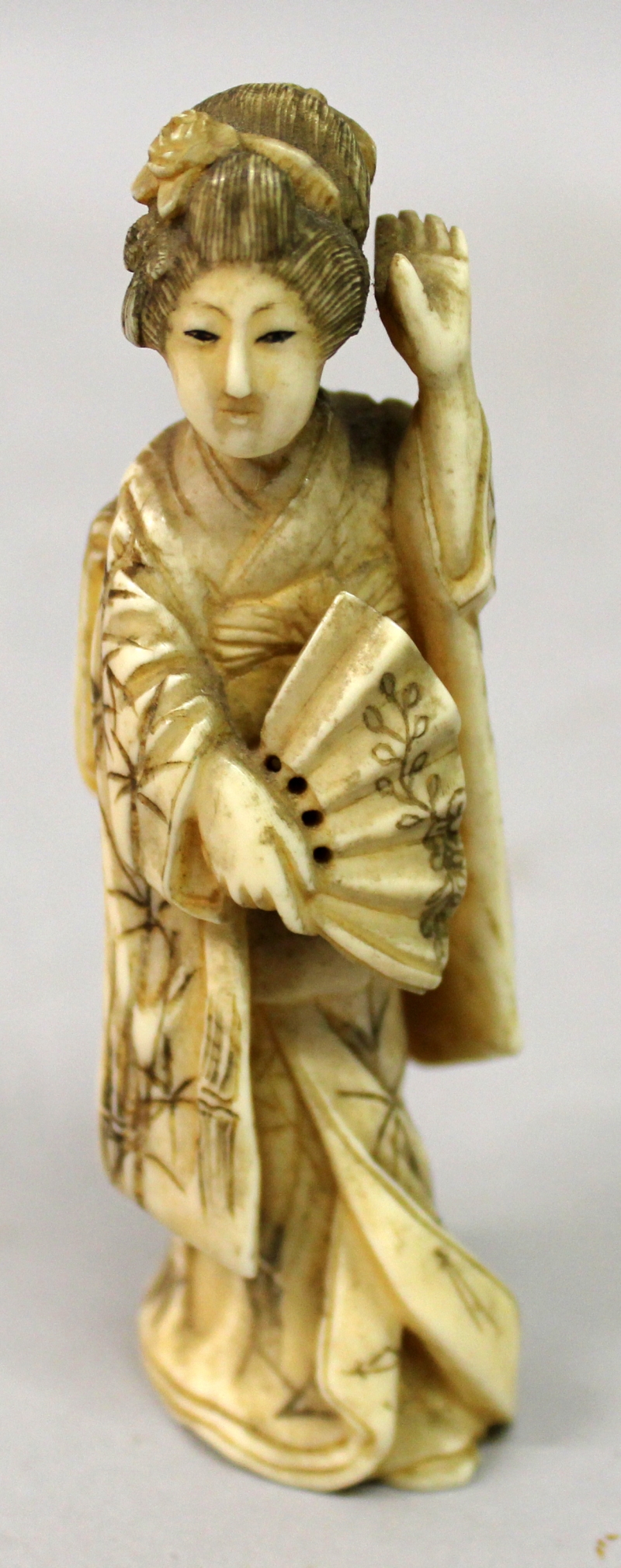 A JAPANESE MEIJI PERIOD IVORY OKIMONO OF A GEISHA, standing in flowing robes and holding a fan,