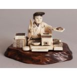 A JAPANESE MEIJI PERIOD SECTIONAL IVORY OKIMONO OF A FOOD SELLER, together with a carved wood stand,