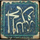 AN IZNIK TILE central script inside a scrolling flower head border on a blue ground. 20cm square (