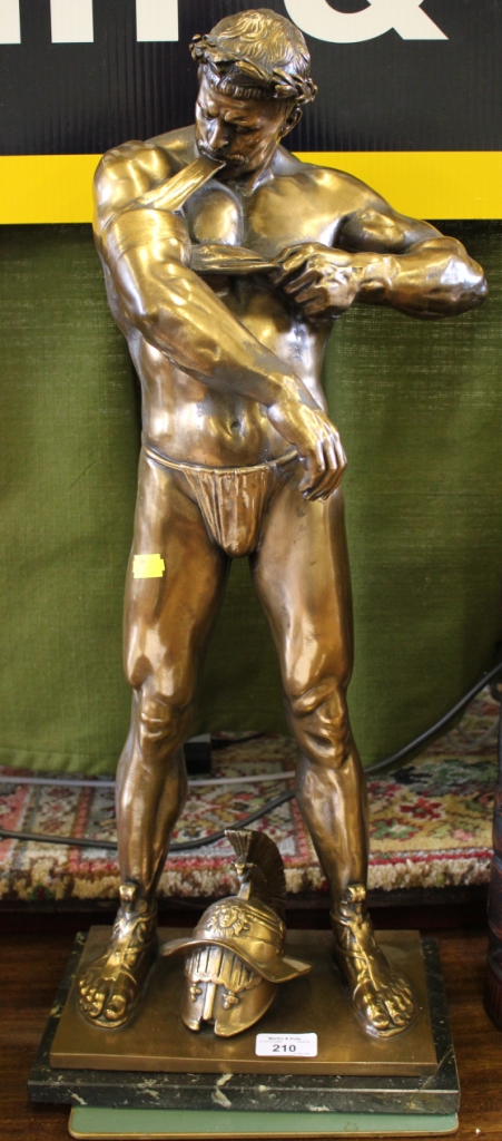 A bronzed metal figure of a gladiator applying a torniquet to his arm, on a marble table 64cm high