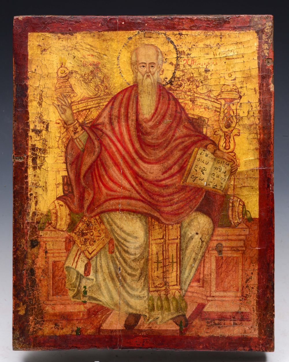 AN ORTHODOX RED AND GILT PAINTED ICON portraying a seated saint holding a Bible, perhaps St Paul,