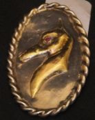 An oval white metal brooch with gilt relief decoration of a greyhound with red stone eye