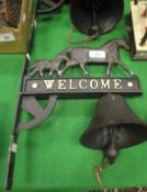 A cast iron bell mounted on a cast iron wall bracket decorated with horses and inscribed "Welcome",