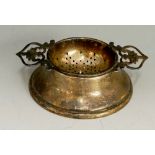 A silver tea strainer and stand, conventional bowl with two pierced handles,