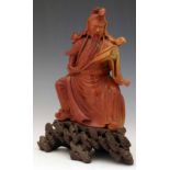 Chinese red soapstone figure of the seated Guandi on a rocky outcrop, reading a scroll, height 24cm.