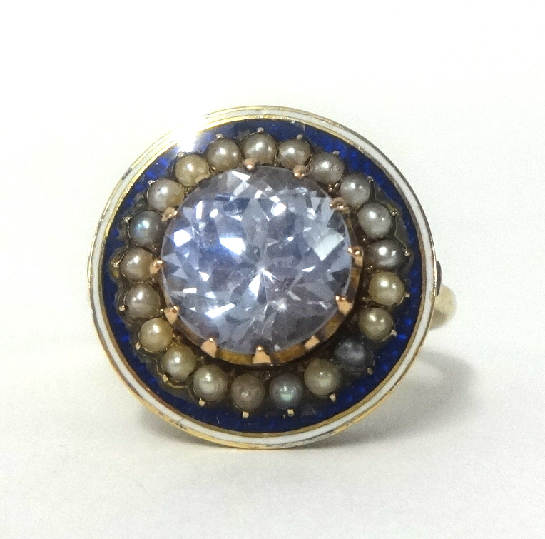 A large pale sapphire ring set within a band of seed pearls and blue enamel, set in a yellow metal