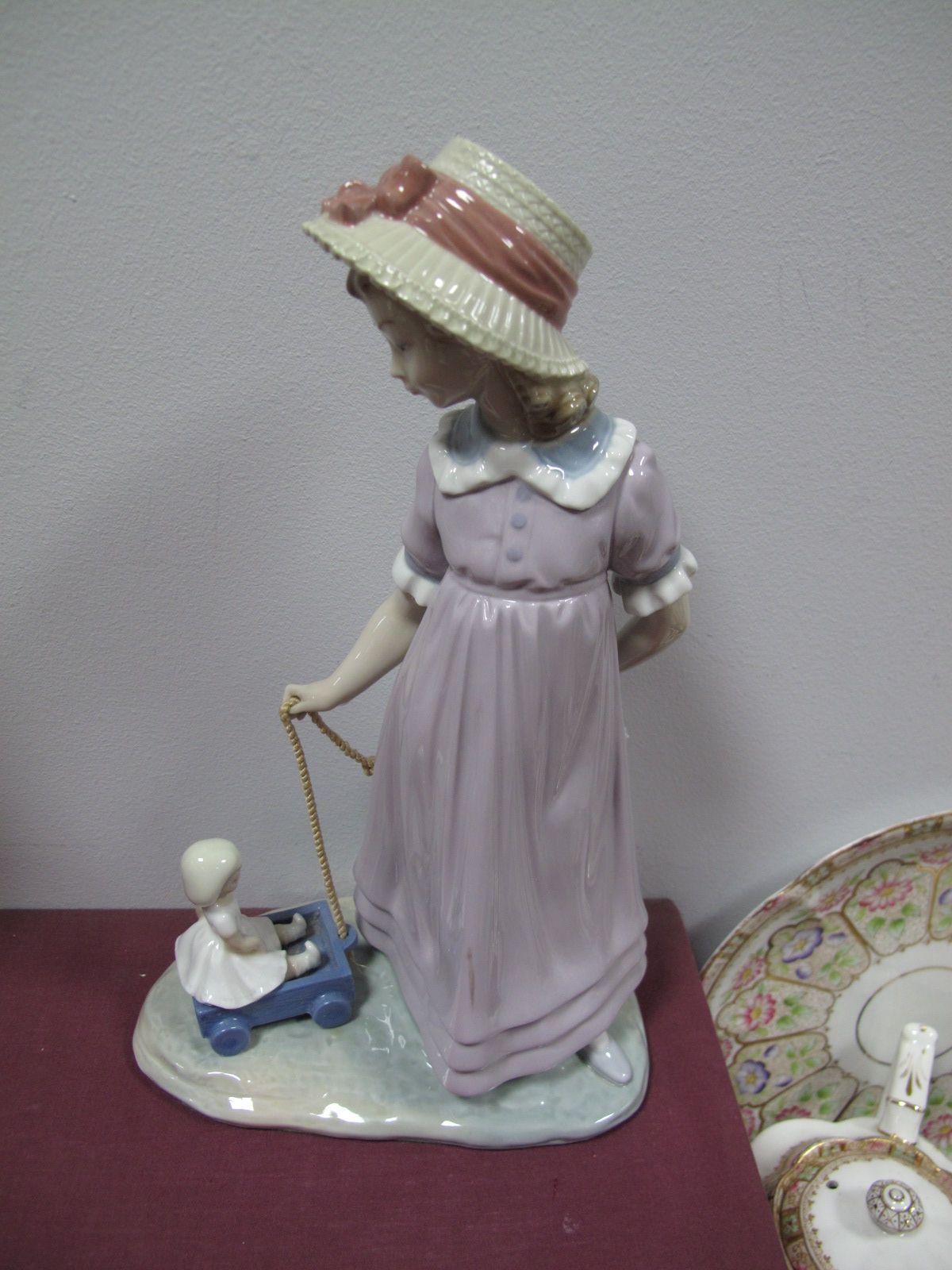 A Lladro Model of a Young Girl Wearing a Bonnet Pulling a Doll on a Kart, all on naturalistic