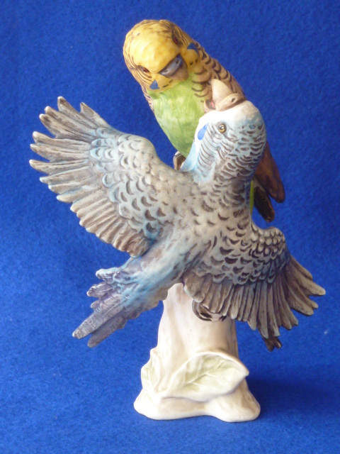 A porcelain Model of a pair of playful Budgerigars on a tree stump decorated in matt polychrome,
