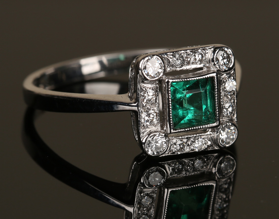 An emerald and diamond ring, the square cut emerald in an openwork surround, mounted with circular