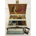 A cream jewellery box containing a quantity of costume jewellery to include beads, brooches, a