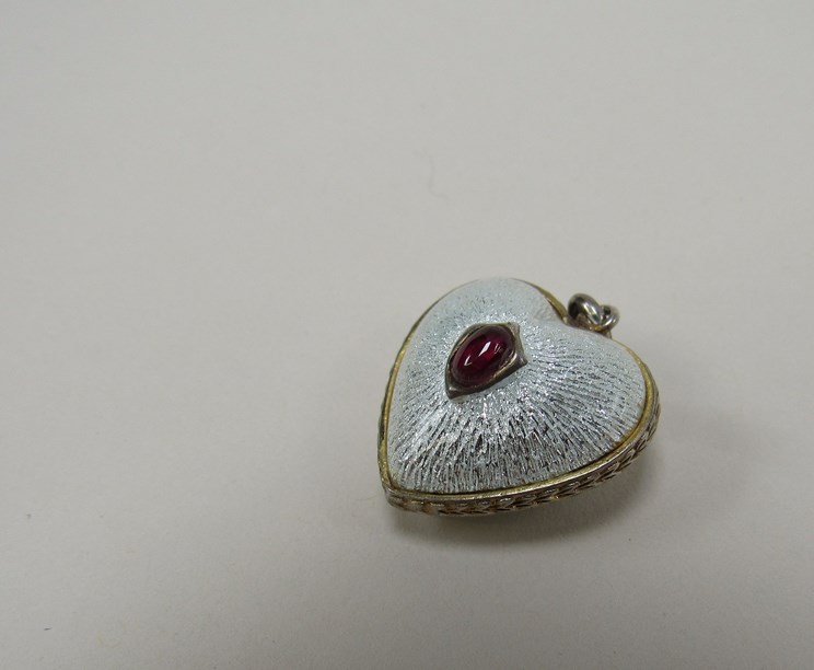 A white guilloche heart shaped pendant with cabochon ruby set to centre