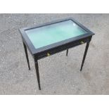 EBONISED DISPLAY TABLE WITH A GLAZED HINGED RISING SLOPING TOP AND A DRAWER, ON TURNED TAPERING LEGS