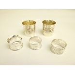 THREE SILVER NAPKIN RINGS TOGETHER WITH A PAIR OF SMALL WHITE METAL BEAKERS, EACH WITH EMBOSSED