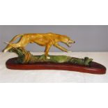 A ceramic model of a greyhound, with a naturalistic base, and raised on a shaped wooden plinth.