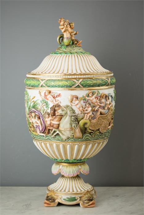 An Italian Capodimonte urn and cover profusely decorated with relief modelled cherubs, chariots,