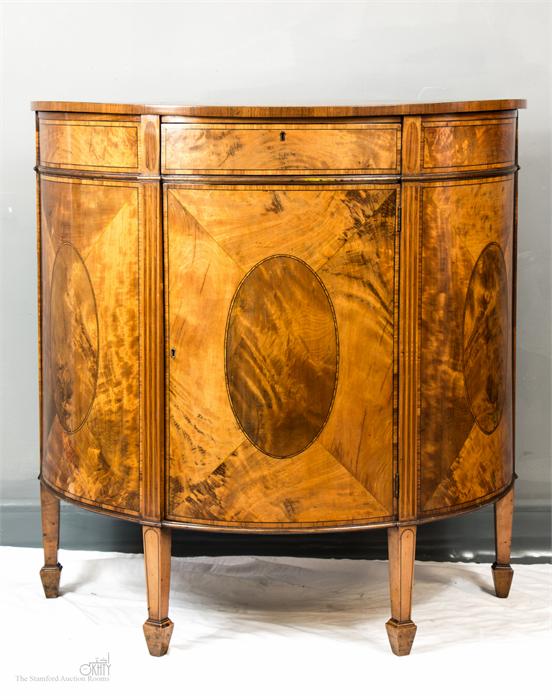 A 19th century demi-lune satinwood and mahogany commode with oval veneered sides and door, raised on