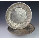 Two Indian white metal circular serving dishes, shaped rims with engraved foliate detailing,