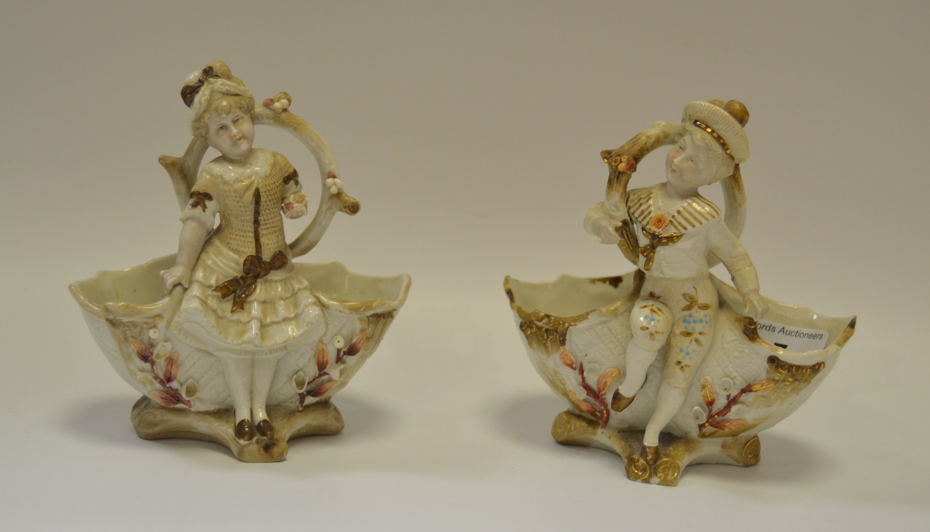 A pair of Continental porcelain figural table salts as Dandy and Beau beneath branches,